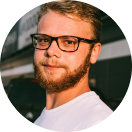 A man with short blond hair, a beard, and black glasses is wearing a white shirt and standing outdoors, looking at the camera.